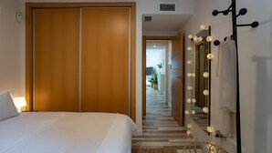 A hotel room with a bed, a wardrobe, a mirror with lights, and a hallway leading to another room.