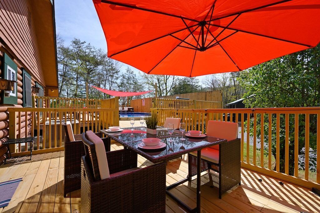 Une terrasse extérieure avec une table dressée pour le repas, entourée de chaises en osier, et un parasol orange offrant de l’ombre.