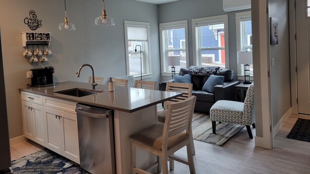 A modern kitchen with a central island, a dining area with a sofa and chairs, and large windows providing views of the outside. - Downtown Charlevoix, Riverwalk 107 (Charlevoix)