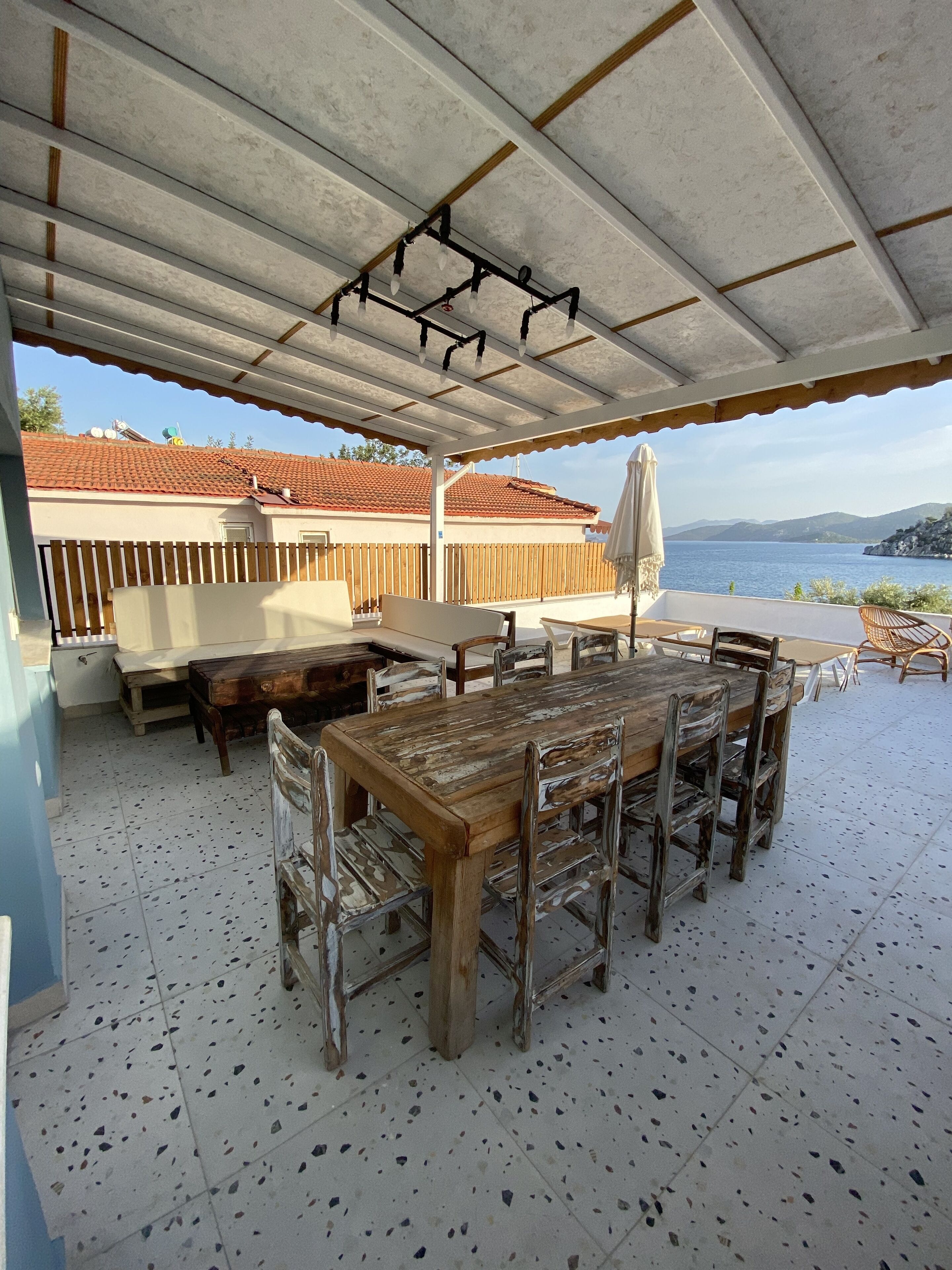 An outdoor patio with a wooden table and chairs, a covered area with a view of the sea and mountains.