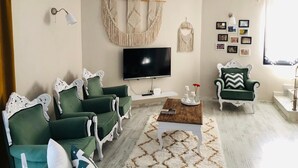 Een woonkamer met een flatscreen televisie, groene gestoffeerde banken, een salontafel, macramé wanddecoraties en een kroonluchter.