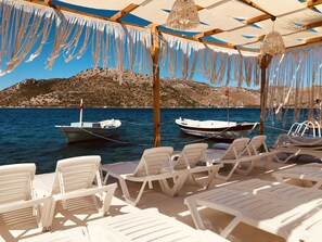 A beachside seating area with white plastic chairs and tables, two boats moored in the water, and a rocky hillside in the background. - Seyyah Bozburun House (Marmaris)