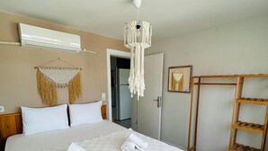 Een net opgemaakt bed met opgerolde handdoeken, een macramé wanddecoratie, een houten plankunit en een aan het plafond bevestigde airconditioning.'