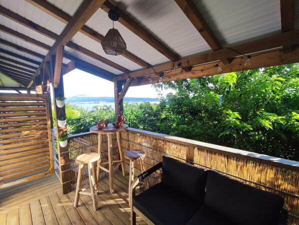 A wooden deck with a black sofa and two stools, a hanging light fixture, and a view of greenery and a body of water in the background.