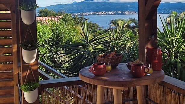 A wooden deck with a table set for breakfast, overlooking a coastal city and mountains. - A little corner of paradise (Les Trois-Îlets)
