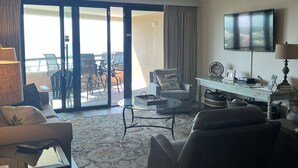 A spacious living room with a large sliding glass door leading to an outdoor area, a flat-screen TV mounted on the wall, and a comfortable seating arrangement including a sofa and two armchairs. - GORGEOUS BEACHFRONT VIEWS!! FREE BEACH SET UP, PRIVATE BEACH, POOL, SPA, AND GYM (Miramar Beach)