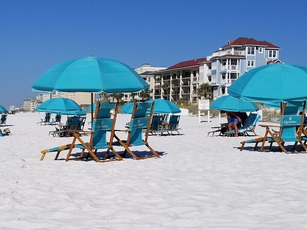 Una playa con hileras de sombrillas azules y sillas de descanso, un edificio con tejado rojo al fondo y un cartel que indica una zona de playa privada.