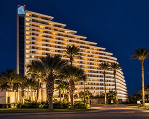 Un hotel de varios pisos con fachada curva, ventanas iluminadas y palmeras en primer plano.