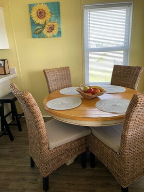 A round dining table with woven chairs, a fruit basket, and a sunflower painting on the wall.