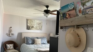 A bedroom with a bed, a ceiling fan, a wall-mounted TV, a wicker chair, and a hat hanging on the wall. - 5 minute walk to beach - come relax, enjoy family time and even fish. (Port St. Joe)