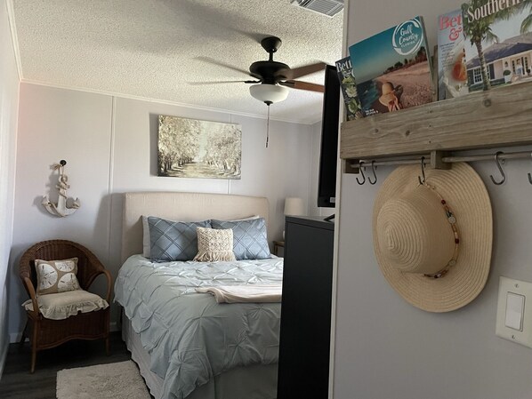 Un dormitorio con una cama, ventilador de techo, televisor montado en la pared, una silla de mimbre y un sombrero colgado en la pared.