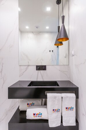 Un baño moderno con pared de mármol, un lavamanos negro y un espejo encima. Hay tres toallas con la marca 'Music Guest House'.