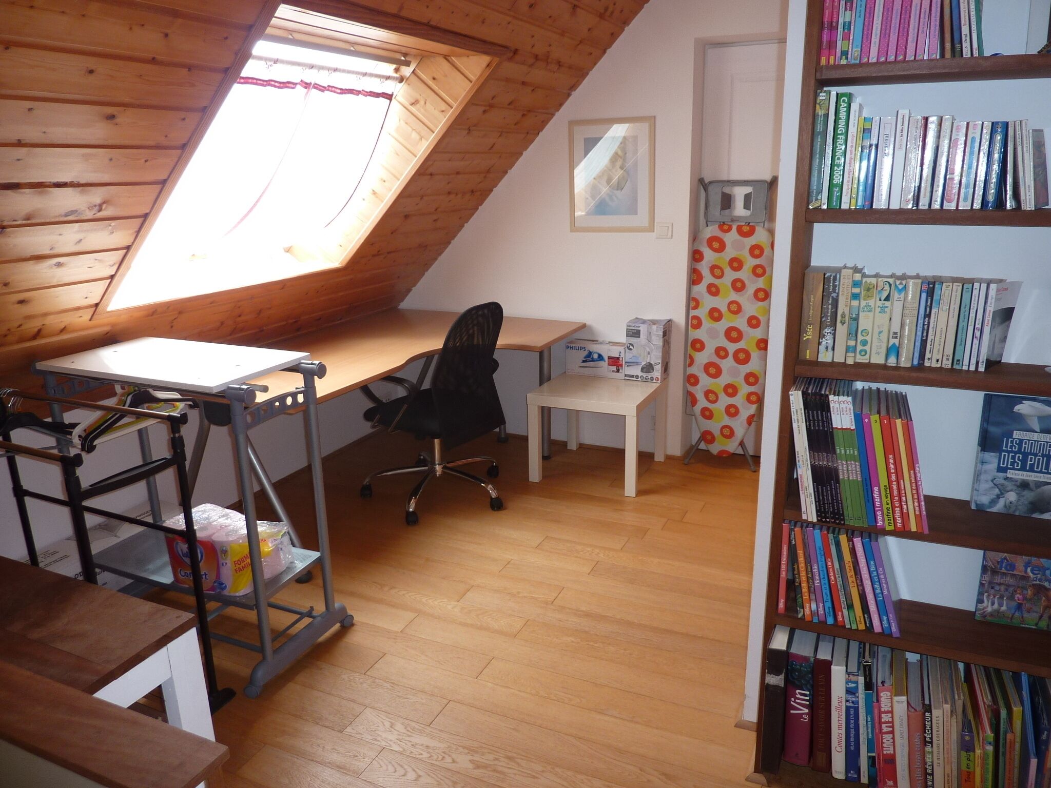 Ein kleines Schlafzimmer unter der Schräge mit Schreibtisch, Stuhl, Bücherregal und einem Fenster mit Vorhang.