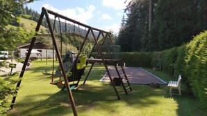 Ein Spielplatz mit Schaukeln und Trampolin, umgeben von Rasen, mit einem Haus im Hintergrund.