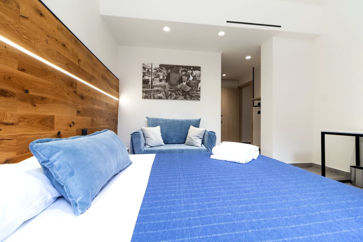 A modern hotel room with a wooden headboard, a large bed with blue bedding, a blue sofa, and a black and white photograph on the wall.