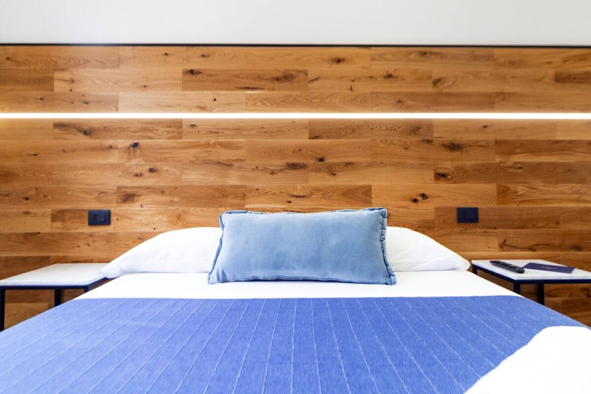 A hotel room with a wooden headboard, a bed with a blue-striped bedspread, white pillows, and a blue pillow. There is a nightstand with a remote control and a book.