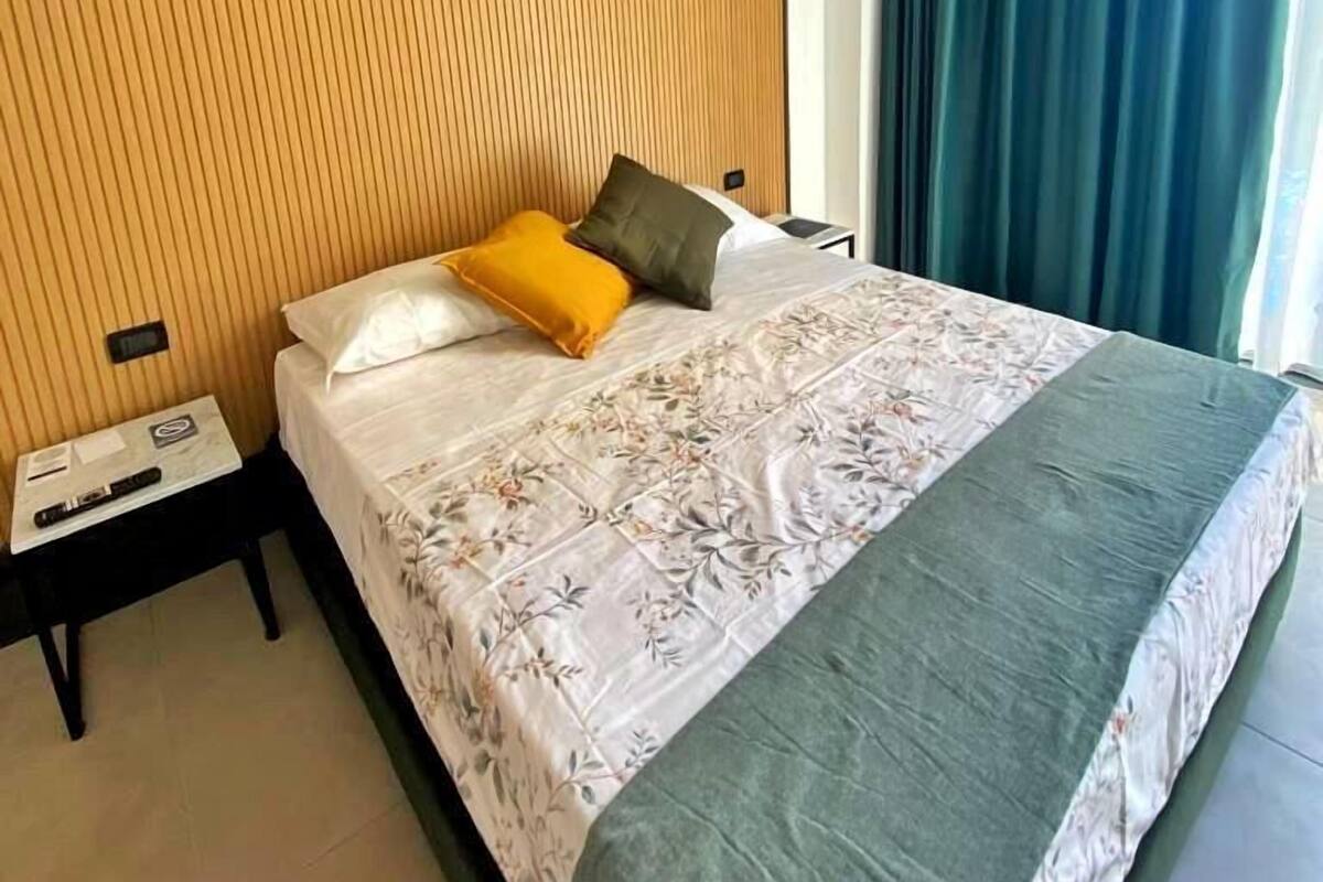 A modern hotel room with a large bed, a wooden headboard, a nightstand, and a window with curtains.