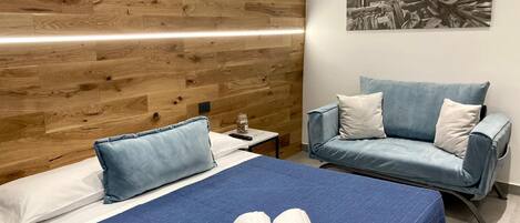 A hotel room with a wooden headboard, a bed with blue bedding, a blue sofa, and a wall-mounted picture.