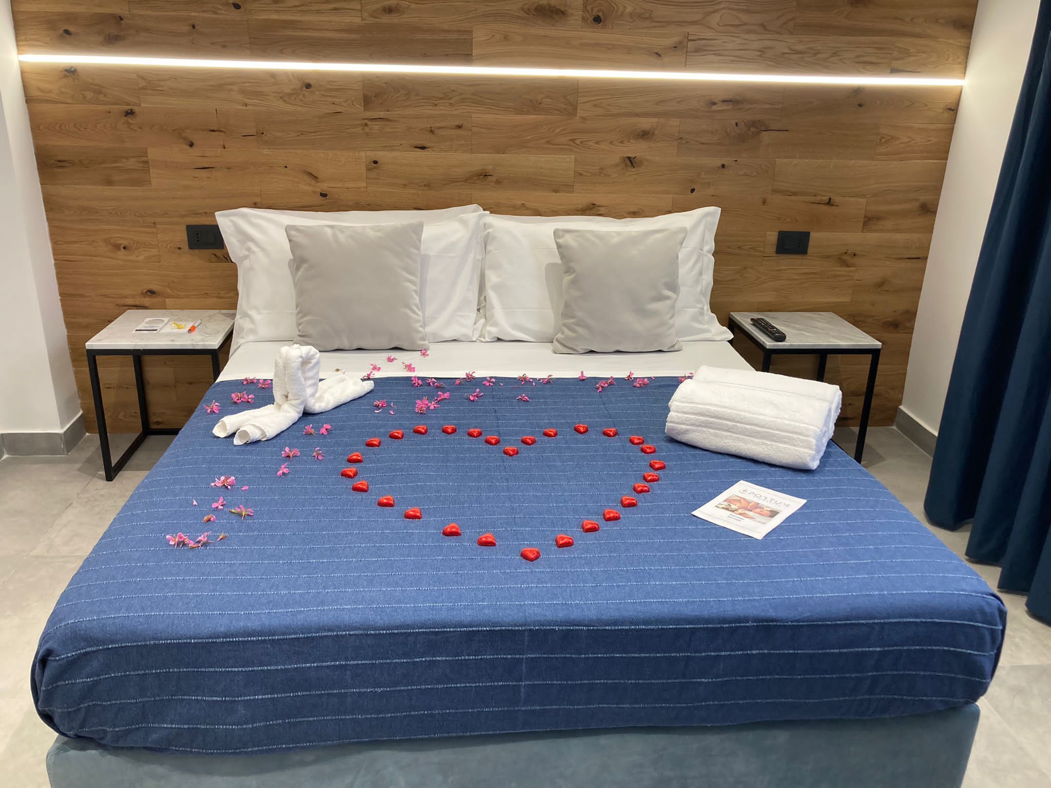 A neatly made bed with a blue bedspread, white pillows, and a heart-shaped decoration made of red flowers and white towels.