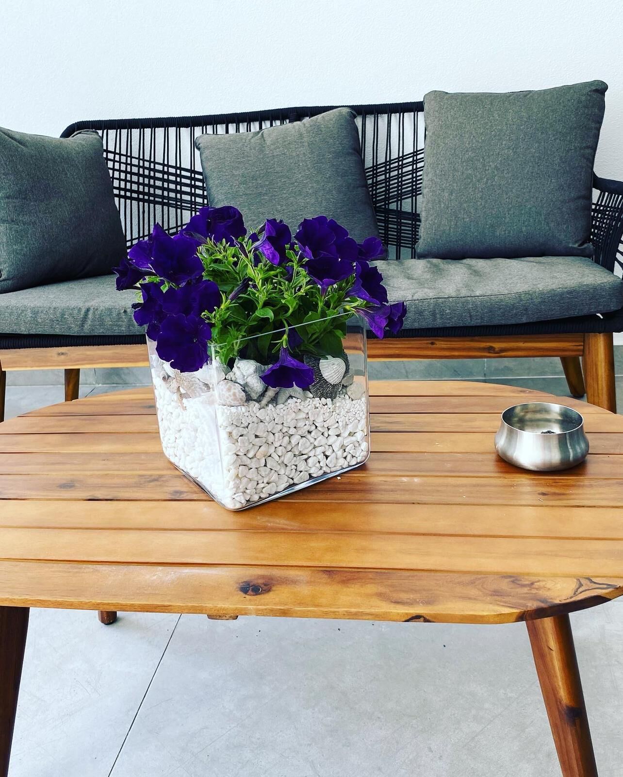 A square glass vase with purple flowers and white pebbles on a wooden table.