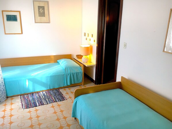 A room with two single beds, a small table with a lamp, and a patterned rug on the floor.