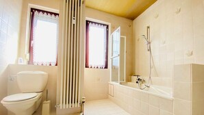 A bathroom with a toilet, a radiator, a shower, and two windows with lace curtains. - Apartment Huningue (Huningue)