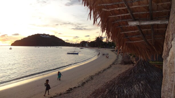 A beach with a thatched-roof structure, calm sea, and a few people walking along the shore.