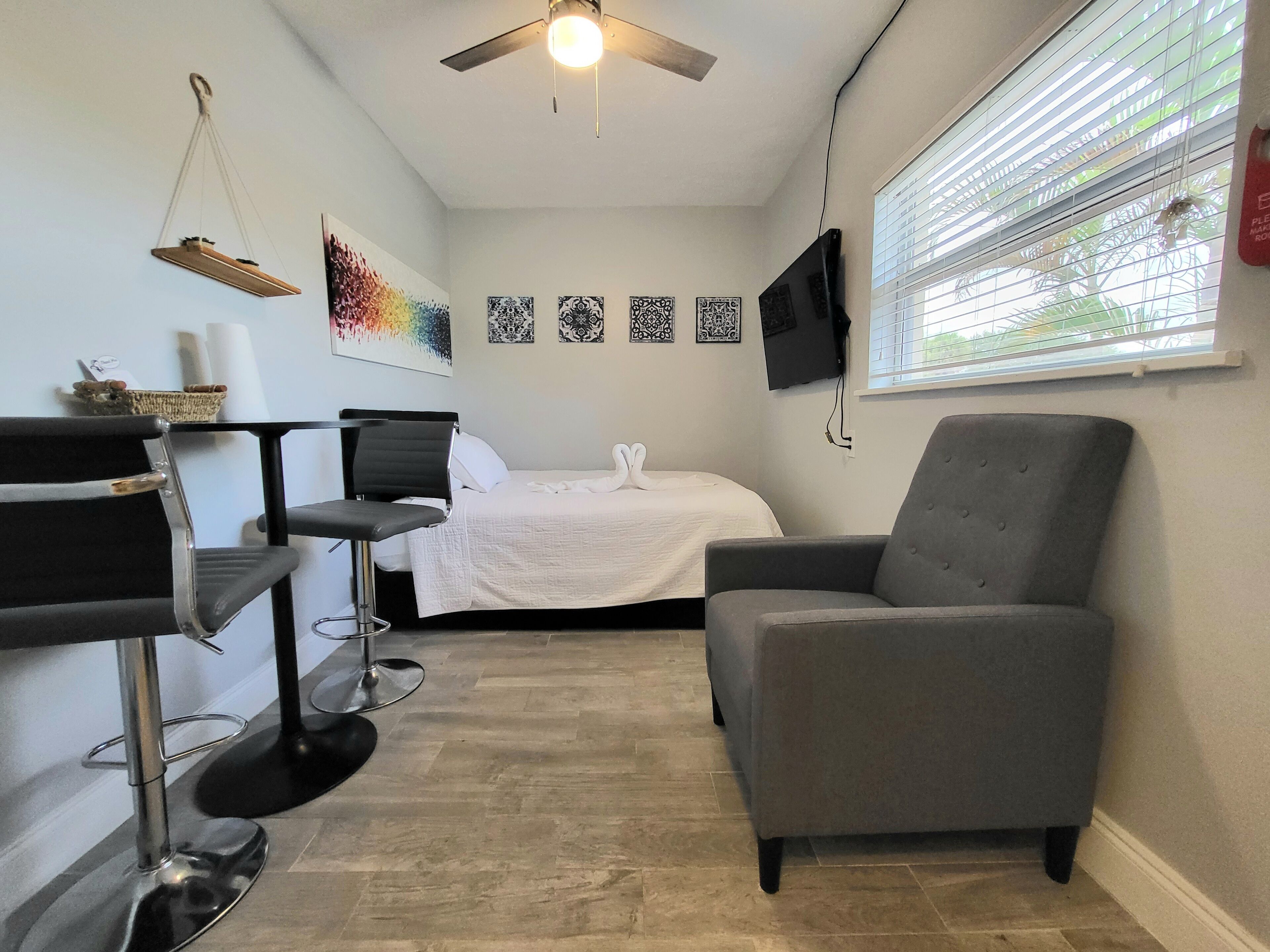 A compact hotel room with a bed, a grey armchair, a small table with a chair, a ceiling fan, and a TV mounted on the wall.