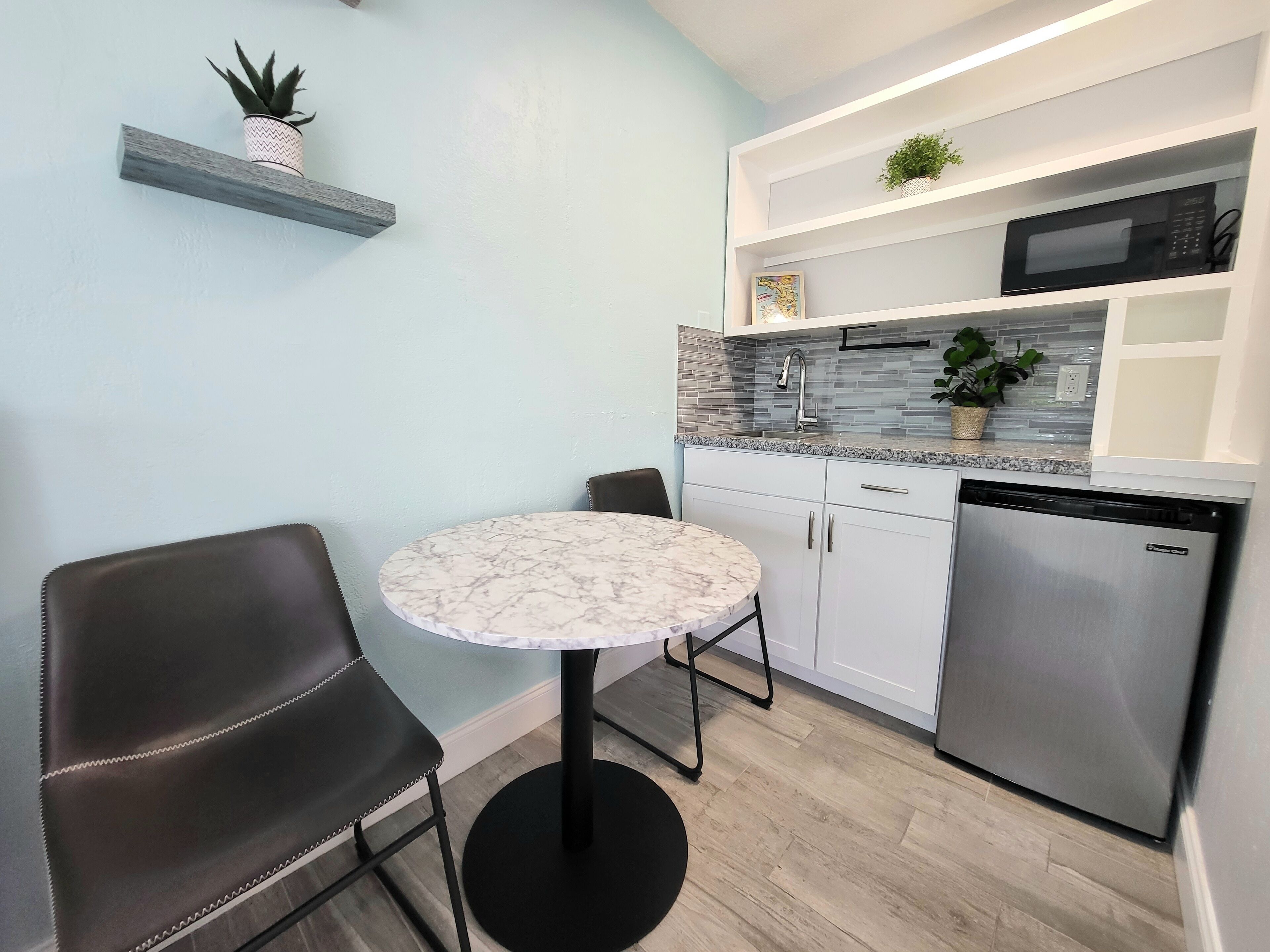 A compact kitchen with white cabinets, a stainless steel dishwasher, a microwave, and a small dining table with two chairs.