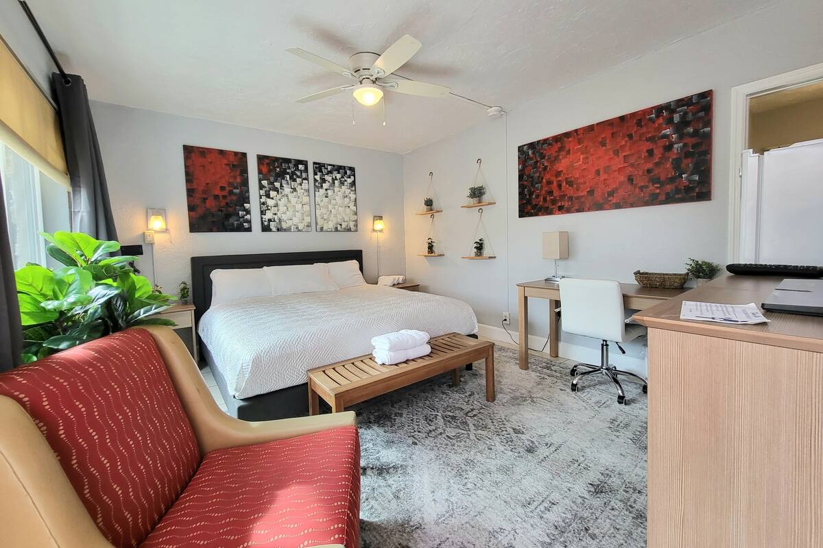 Een hotelkamer met een bed, bureau, stoel, bureaulamp, een klein tafeltje, een plant en kunst aan de muren.