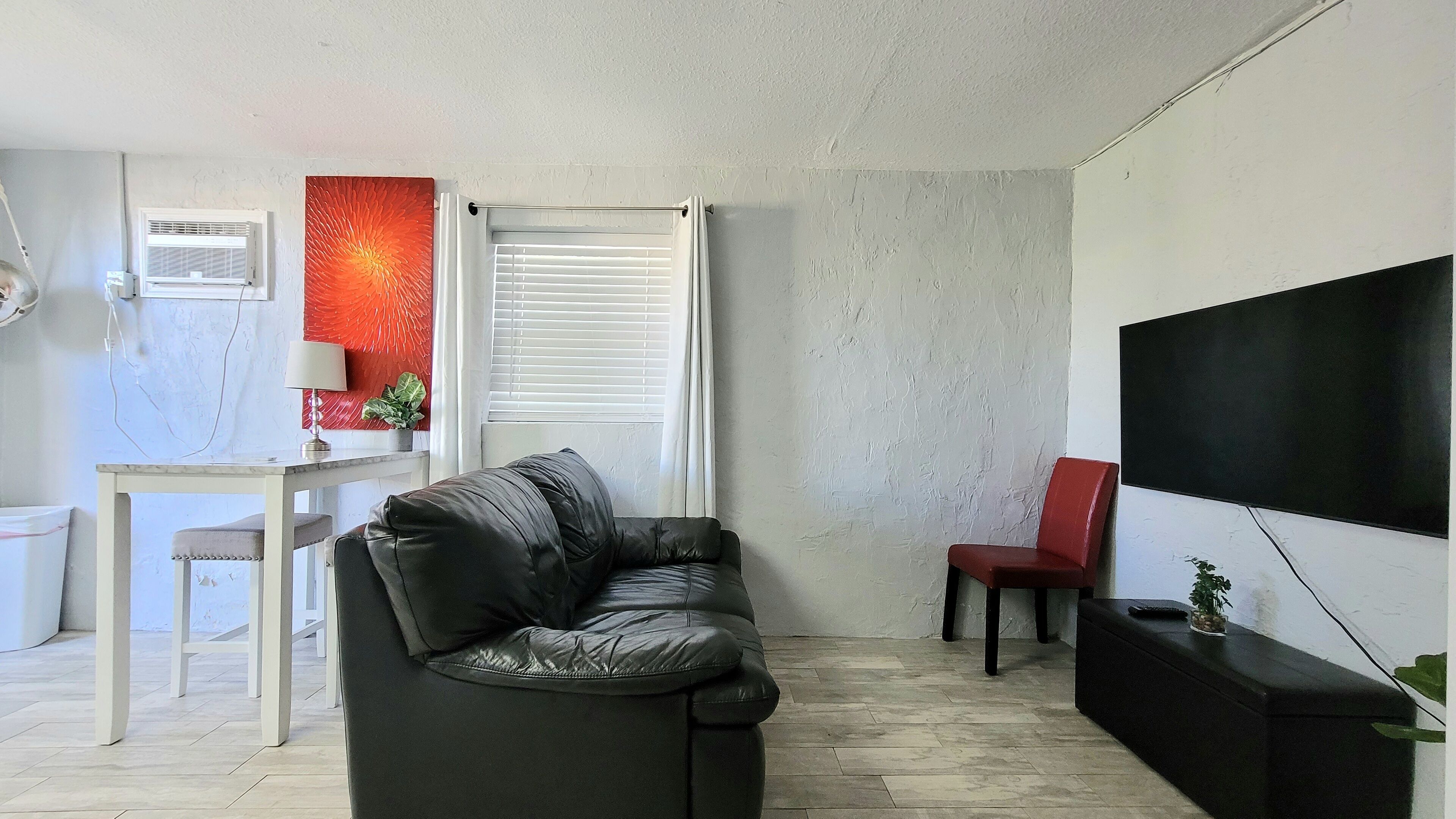 Una sala de estar con un sofá de cuero negro, un sillón rojo, una mesa blanca con una lámpara, un televisor en la pared y una ventana con persianas.