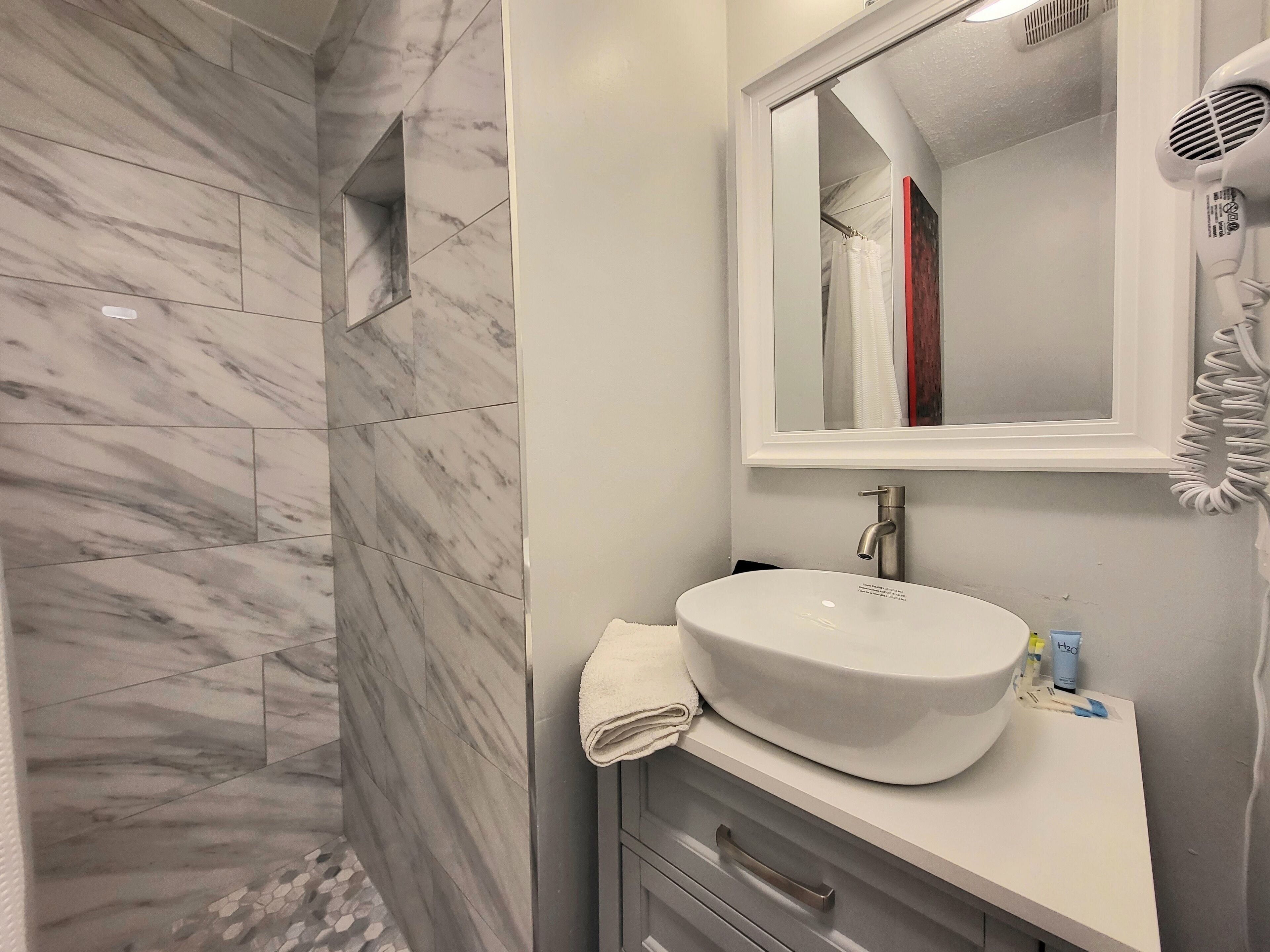 A bathroom with a marble-tiled shower, a white sink, a mirror, and a hairdryer.