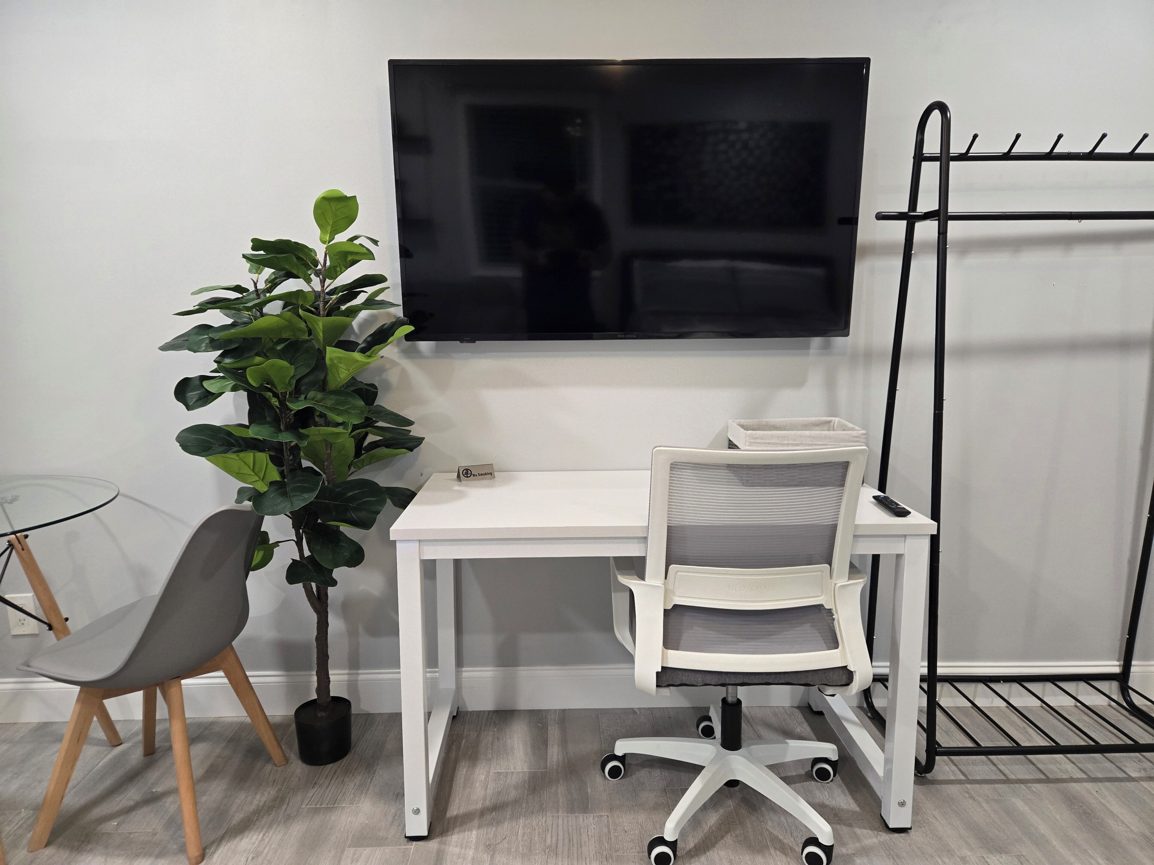 Un espacio de oficina moderno con un escritorio blanco, una silla de oficina gris, una planta en maceta y un televisor montado.