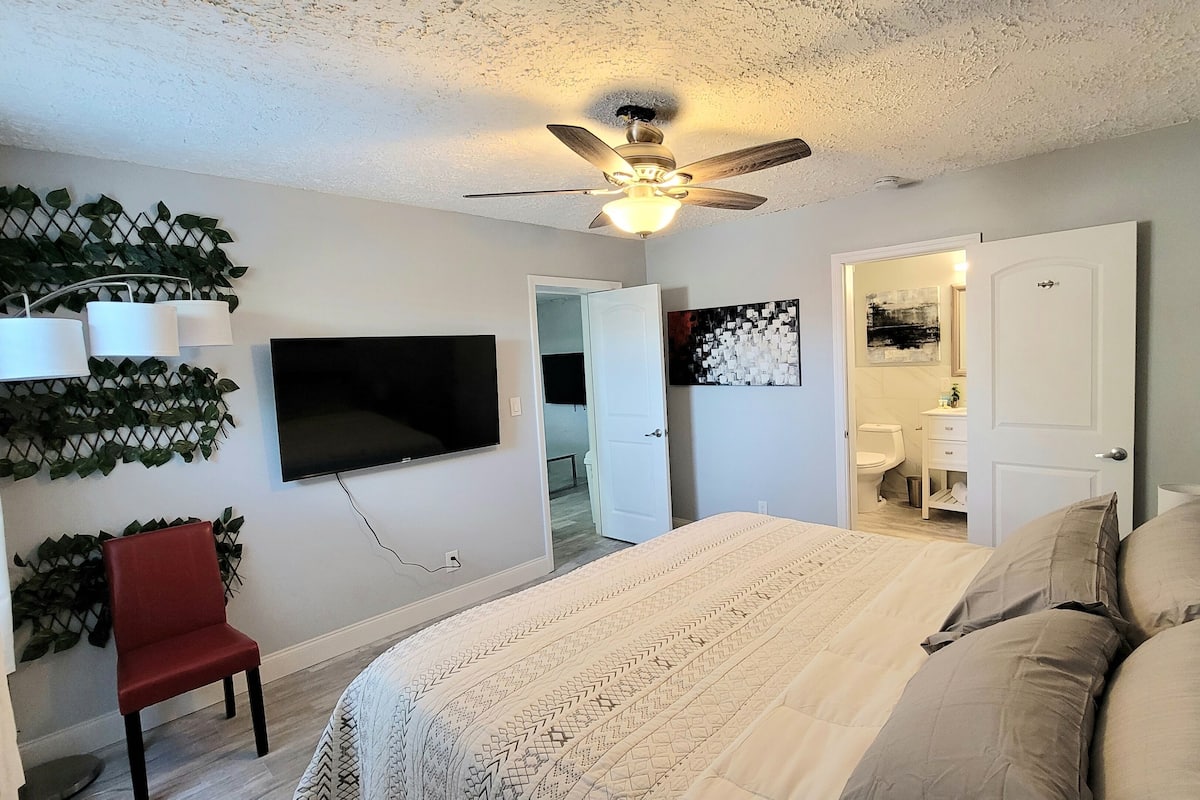 Slaapkamer met een bed, een rode stoel, een plafondventilator, een televisie en een badkamer zichtbaar door een geopende deur.