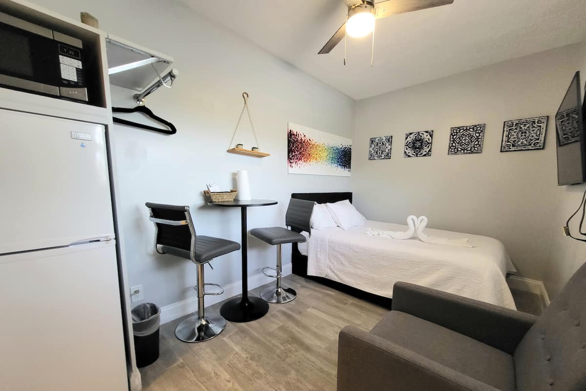 Een kleine hotelkamer met een bed, twee stoelen, een tafeltje, een koelkast en een plafondventilator.
