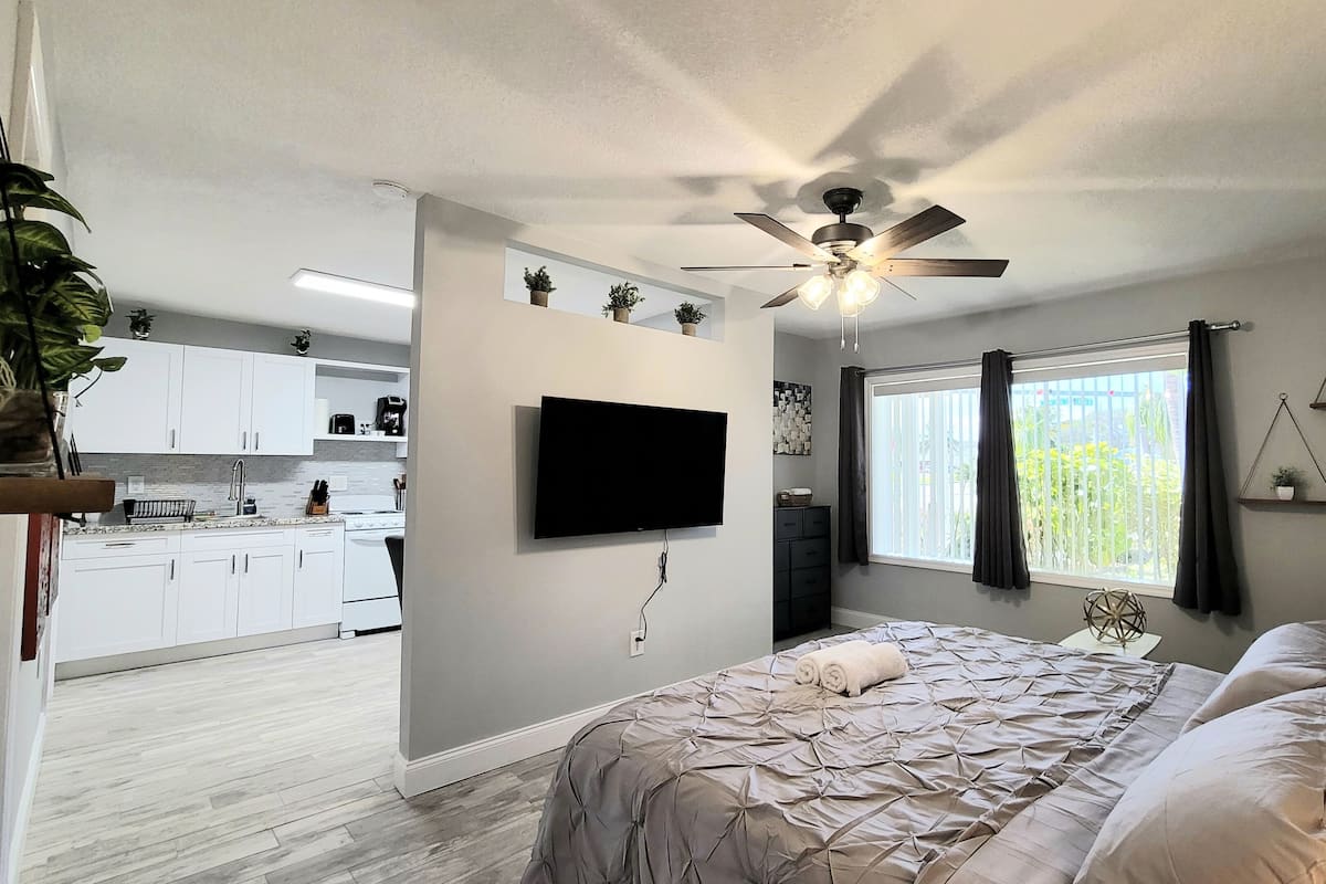 Een slaapkamer met een bed, een plafondventilator, een televisie, een raam met gordijnen en uitzicht op de keuken.
