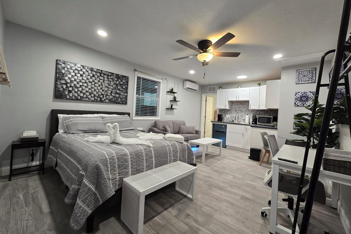 Een moderne slaapkamer met een groot bed, nachtkastjes, een bankje, een plafondventilator en een kitchenette op de achtergrond.