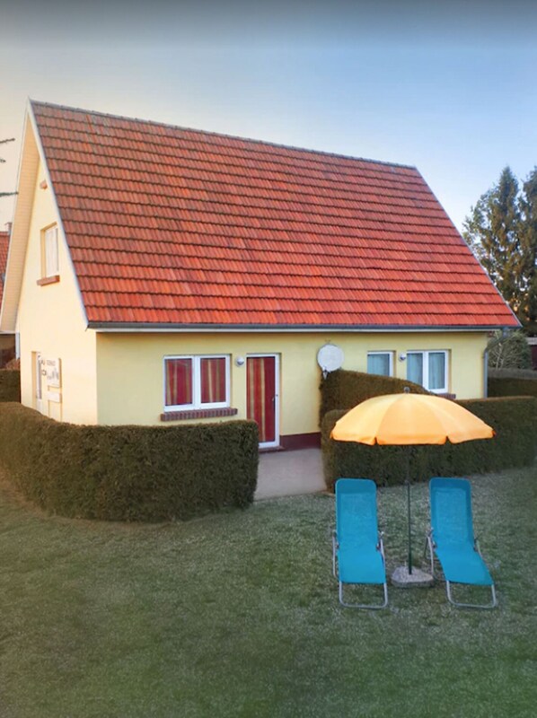 Une maison avec un toit rouge, une porte d’entrée et deux fauteuils bleus placés à l’extérieur, sous un parasol.