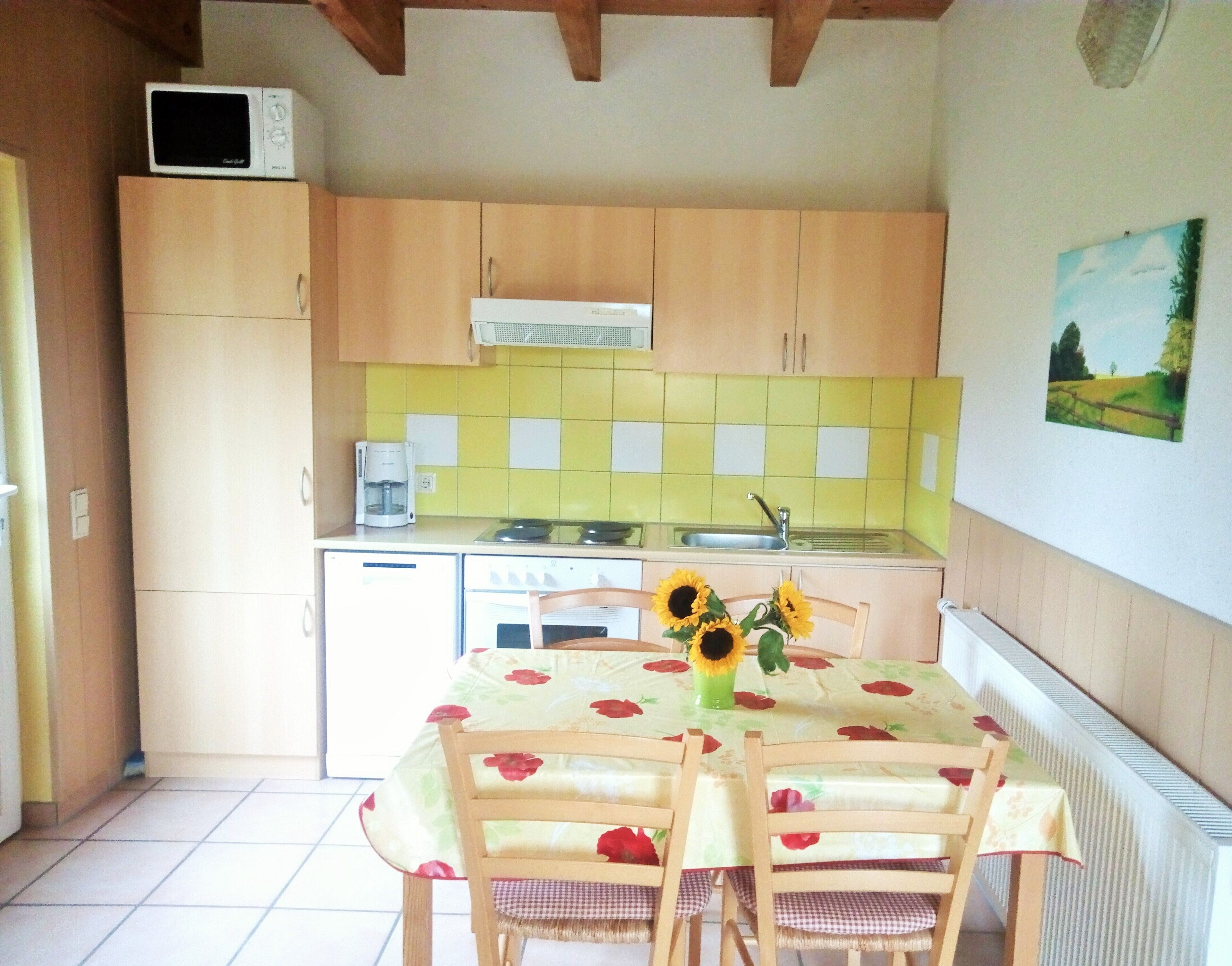 Een kleine keuken met houten kastjes, een magnetron, een koffiezetapparaat en een tafel met stoelen. Er hangt een schilderij aan de muur en een raam met gordijnen.