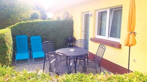 Une terrasse avec une table ronde et quatre chaises, une petite bordure en briques et une fenêtre avec des stores.