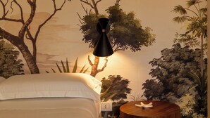 Een slaapkamer met een bed, een rond nachtkastje, een muurschildering van bomen en een lamp op het nachtkastje.