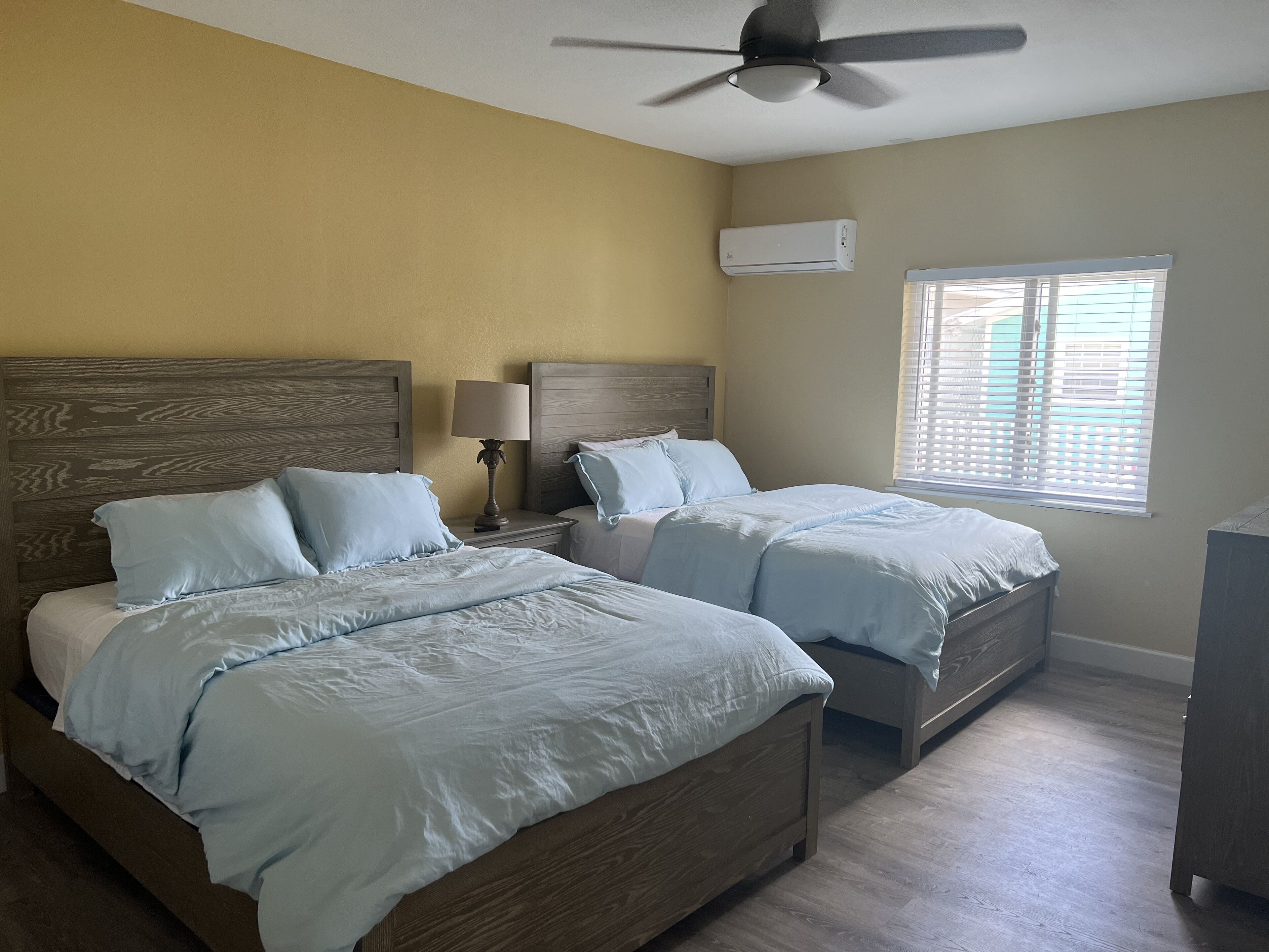 Une chambre avec deux lits, des têtes de lit en bois, un ventilateur de plafond, un appareil de climatisation et une fenêtre avec des stores.