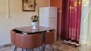 Un espai vital reduït amb una taula rodona i quatre cadires, un refrigerador blanc, una impressió botànica emmarcada a la paret i una cortina vermella que cobreix parcialment una finestra.