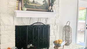 Een open haard met een witte stenen schouw, een lijstwerk landschap schilderij, en decoratieve items, waaronder pompoenen en een mand met fruit.