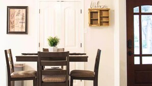 Een eethoek met een houten tafel, vier stoelen, een hangende lamp, een deur met een raam en een wandplank.