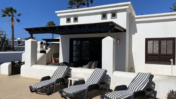 A modern building with a white exterior, a covered patio area with striped lounge chairs, and a large window with dark shutters. - Casa Sophia (Playa Blanca)