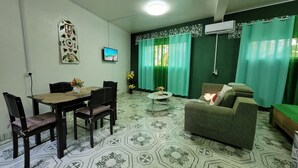 A living room with a sofa, a dining table with chairs, a TV mounted on the wall, and a patterned floor.