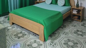 A bedroom with a green bedspread, matching pillows, a wooden headboard, a nightstand with a vase of flowers, and patterned hexagonal floor tiles.