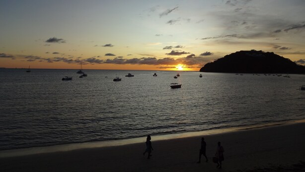 A beach with boats on the water and people walking on the shore.