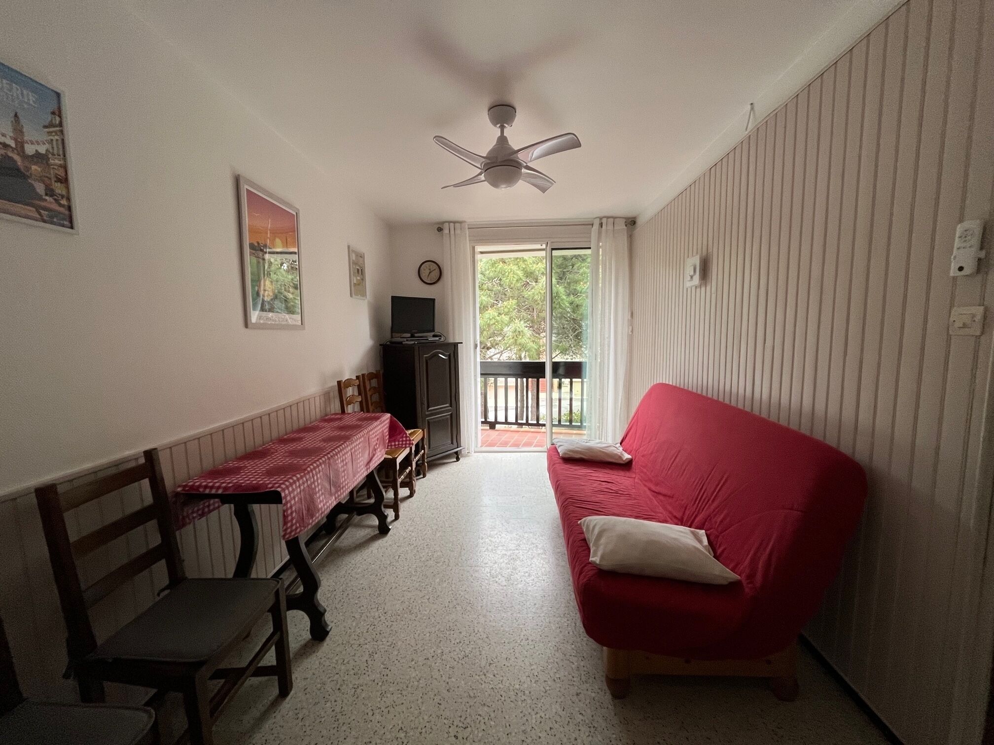 Ein Zimmer mit einem roten Sofa, einem Holztisch mit roter Tischdecke, einem Deckenventilator und Blick ins Freie.